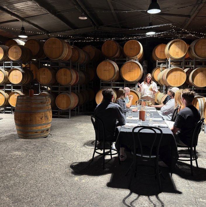 A host leading a seated tasting at a long table in a Hunter Valley barrel hall