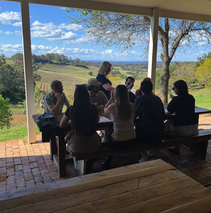 A tour group around a long timber table on a cellar door verandah above the vines